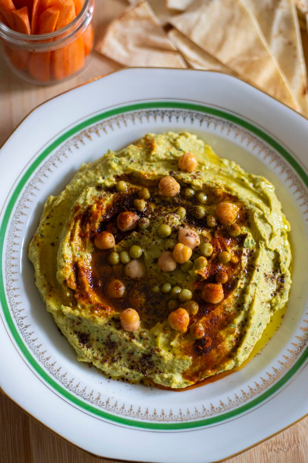 Green Hummus with chickpeas and green peas, paprika and sumac toppings, next to carrot sticks and triangled pita bread