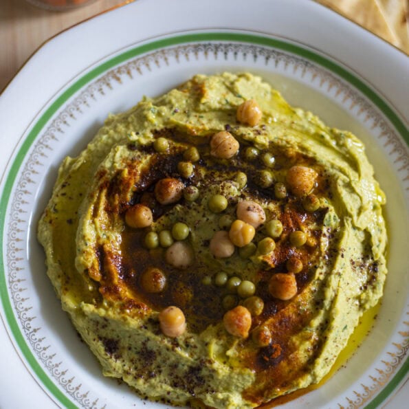 Green Hummus with chickpeas and green peas, paprika and sumac toppings, next to carrot sticks and triangled pita bread