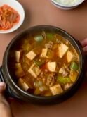 Brown Korean Stew in an earthenware pot, with tofu cubes, beef, zucchini -and sides of kimchi and rice