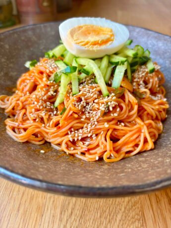 Bibim guksu — Korean spicy cold noodles with gochujang sauce, topped with cucumber matchsticks, sesame seeds and a halved boiled egg, served in a stone bowl