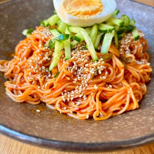 Bibim guksu — Korean spicy cold noodles with gochujang sauce, topped with cucumber matchsticks, sesame seeds and a halved boiled egg, served in a stone bowl