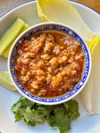 Small bowl filled with nam prik ong, surrounded by crunchy veggies, cilantro, lime