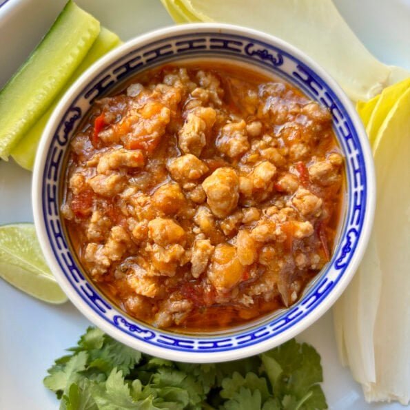 Small bowl filled with nam prik ong, surrounded by crunchy veggies, cilantro, lime